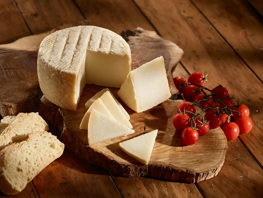 Se muestra un queso partido con una cuña y varios trozos para degustar. Todo sobre una tabla de madera en la que también se muestra unos tomates pequeños.