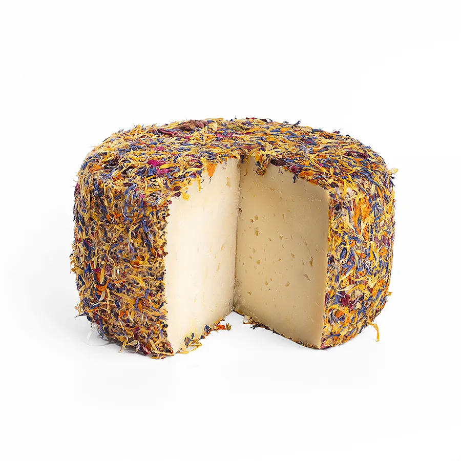 Se muestra un queso entero con una cuña cortada. El queso está cubierto completamente por pétalos de flores, con un aspecto muy colorido.