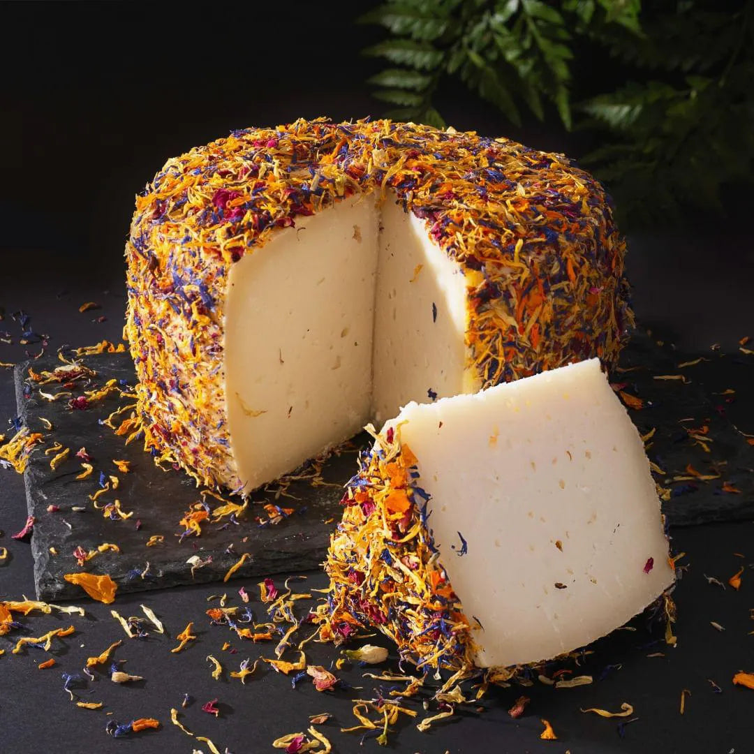 Se muestra un queso entero con una cuña cortada. El queso está cubierto completamente por pétalos de flores, con un aspecto muy colorido.