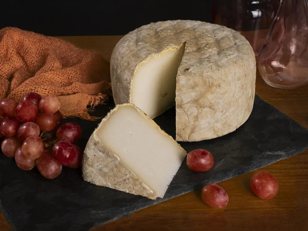 Se muestra un queso entero de corteza gris con una cuña partida. Junto al queso hay unas uvas rojas.