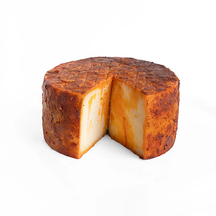 Se muestra un queso cubierto de pimentón sobre un fondo blanco. Al queso se le ha cortado una cuña y se aprecia el interior del queso, que igualmente está manchado de pimentón.