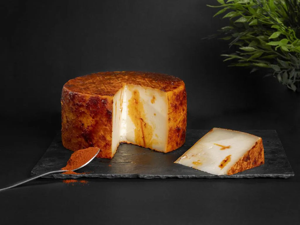 Se muestra un queso entero al que se le ha cortado una cuña. El queso está cubierto de pimentón, y junto al queso hay una cuchara con pimentón.