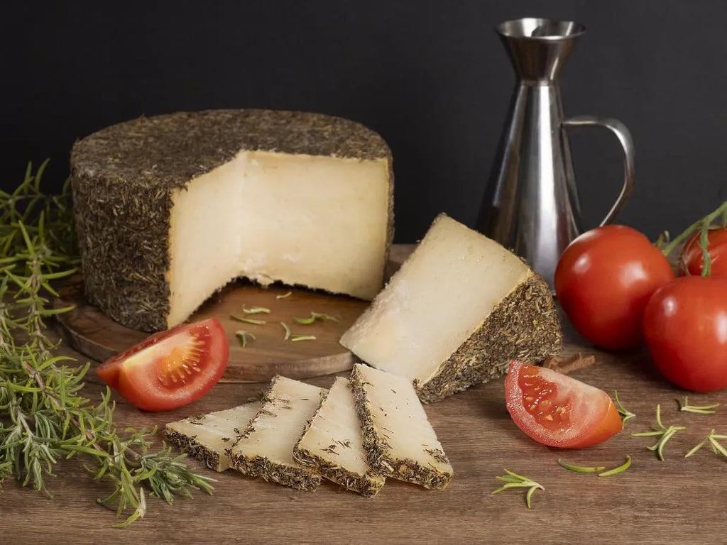 Se muestra un queso entero al que se le ha cortado una gran cuña y se ha partido en pedazos más pequeños. El queso y las cuñas tienen una corteza de tomillo. Junto al queso hay tomates y una jarra de aceite.