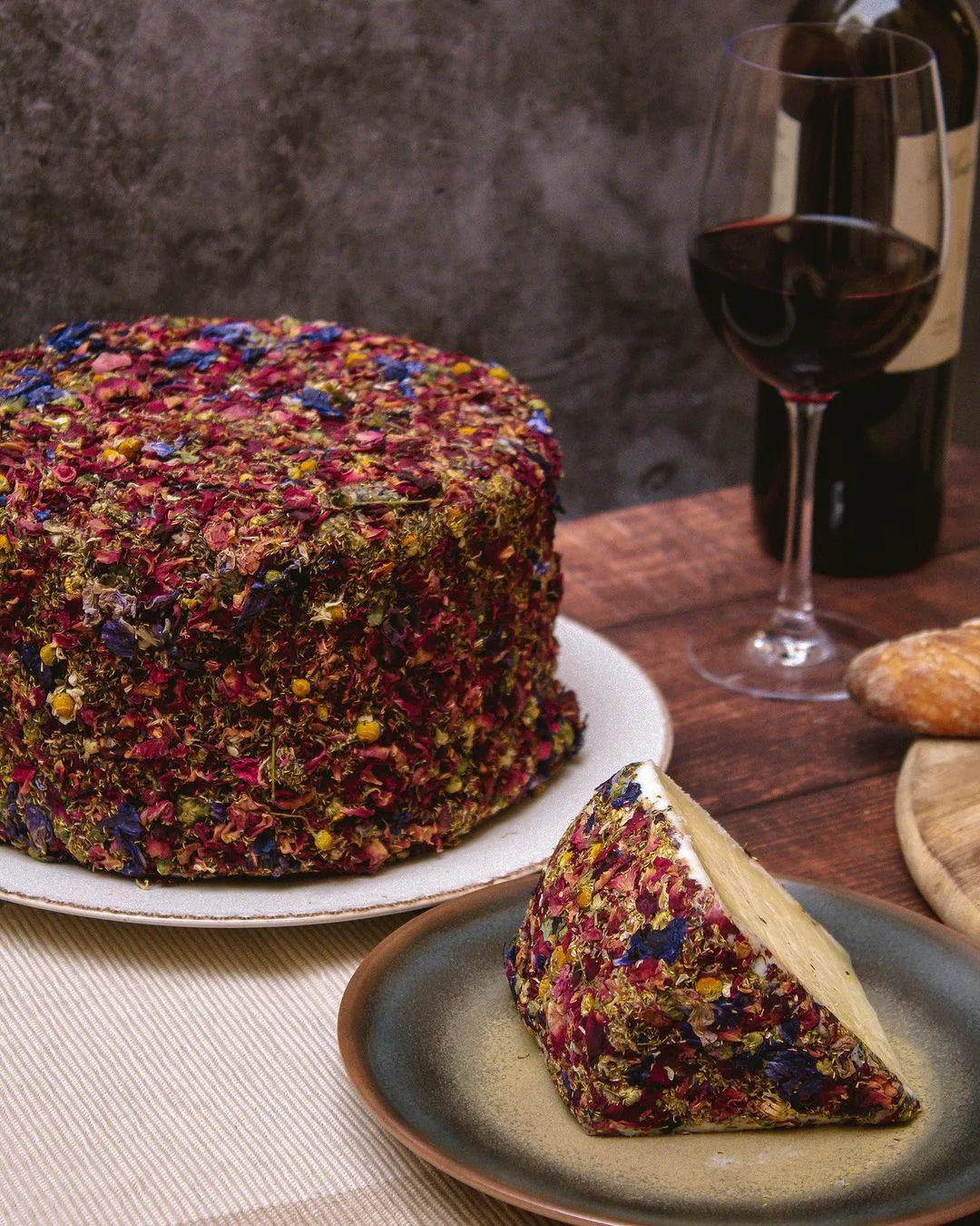 Queso entero sin partir recubierto completamente de pétalos de flores. Al lado se muestra una cuña de dicho queso, y al fondo una copa y una botella de vino tinto.