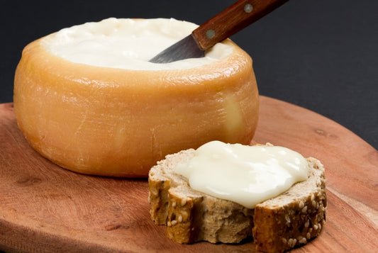 Se muestra un queso estilo torta con muy cremoso, casi líquido. El queso está abierto por arriba y hay una rebanada de pan untada delante del queso.