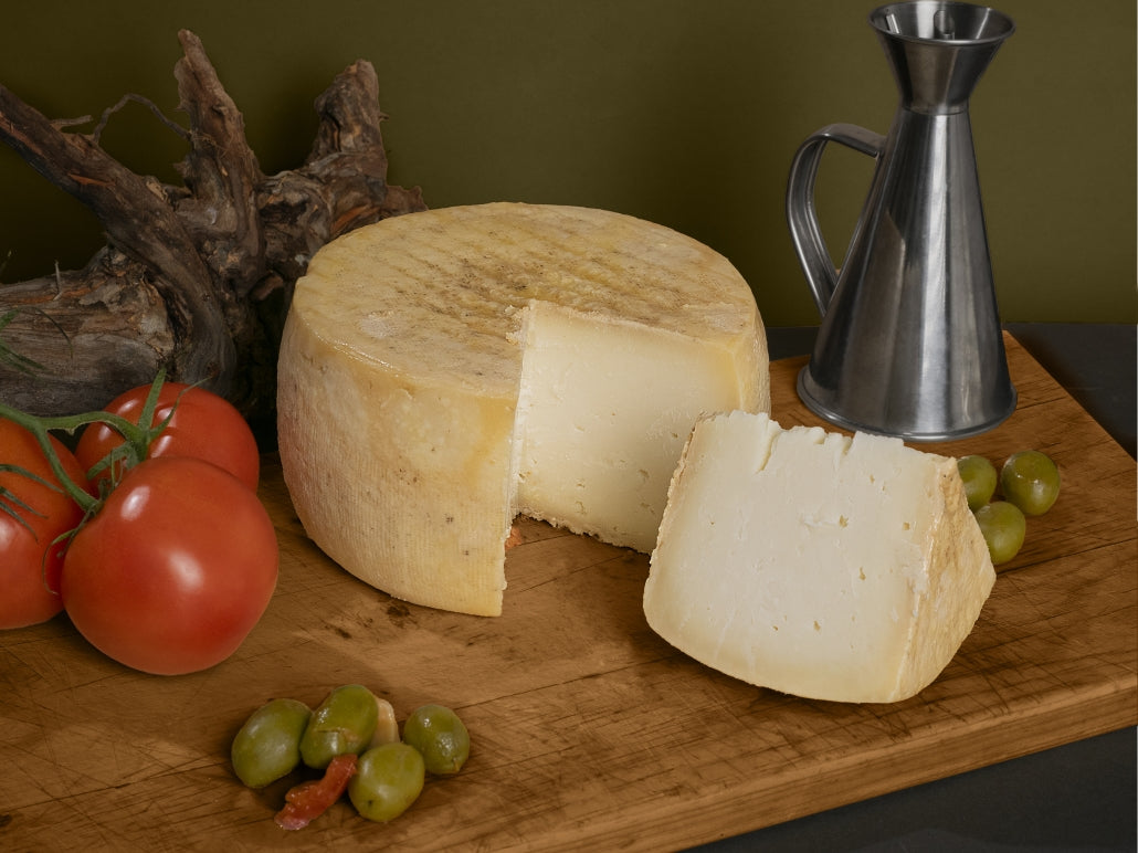 Se muestra un queso entero al que se le ha cortado una cuña y se aprecia su interior blanco. El queso está sobre una tabla con aceitunas, tomate y una jarra de aceite.