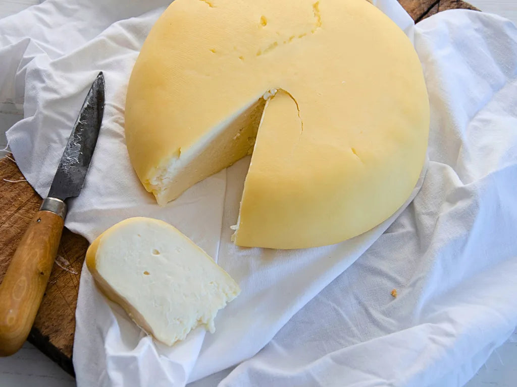 Se muestra un queso de corteza amarilla e interior blanco cremoso. Se muestra el queso entero y una cuña cortada con un cuchillo.
