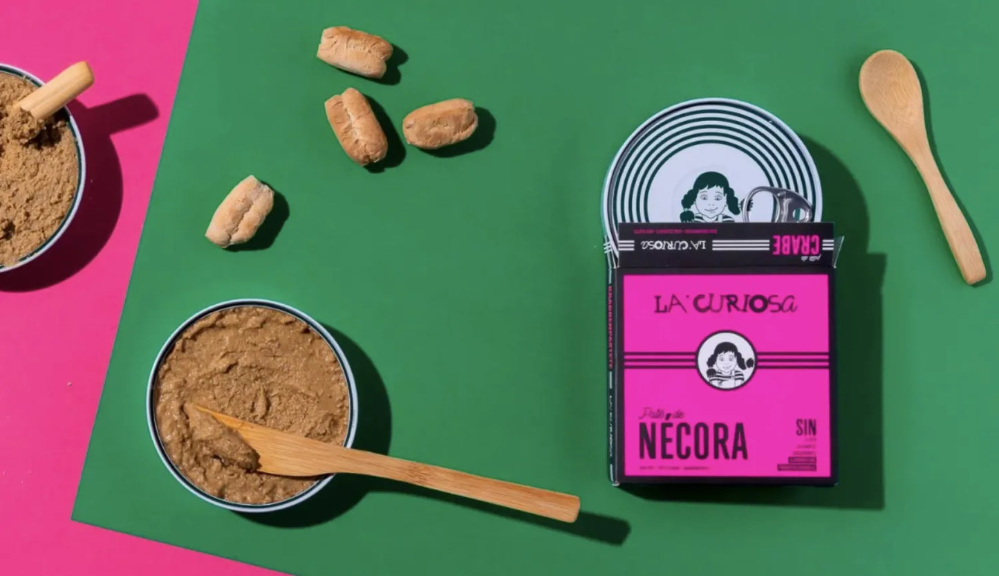 Se muestran varias latas de paté de nécora y varias cajas de color rosa sobre un fondo verde.