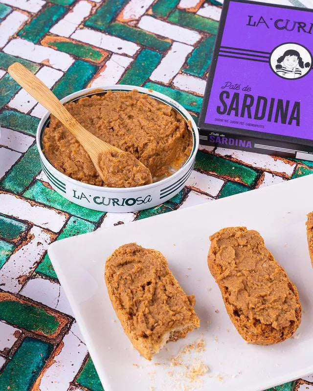 Se muestra una lata de paté  de sardina con su caja de color morado. De la lata se han untado dos tostas y una de ellas tiene un bocado. 