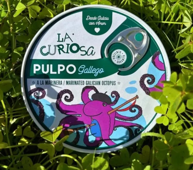 Se muestra la tapa de una lata de pulpo a la gallega con un pulpo morado. La lata está sobre un césped verde.