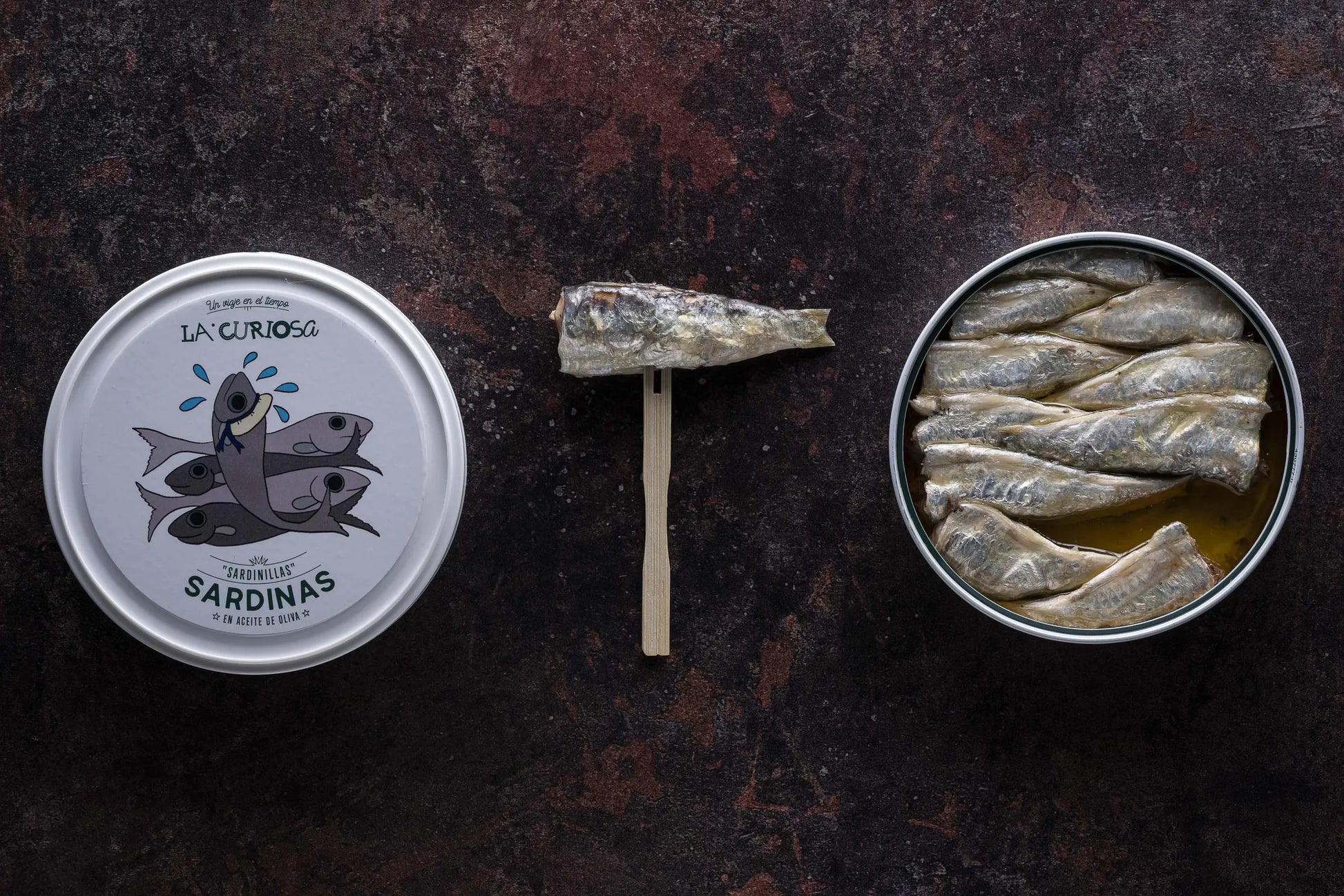 Se muestra una lata de sardinillas con una sardinilla fuera de la lata pinchada en un tenedor de madera. La tapa de la lata está junto a la lata. 