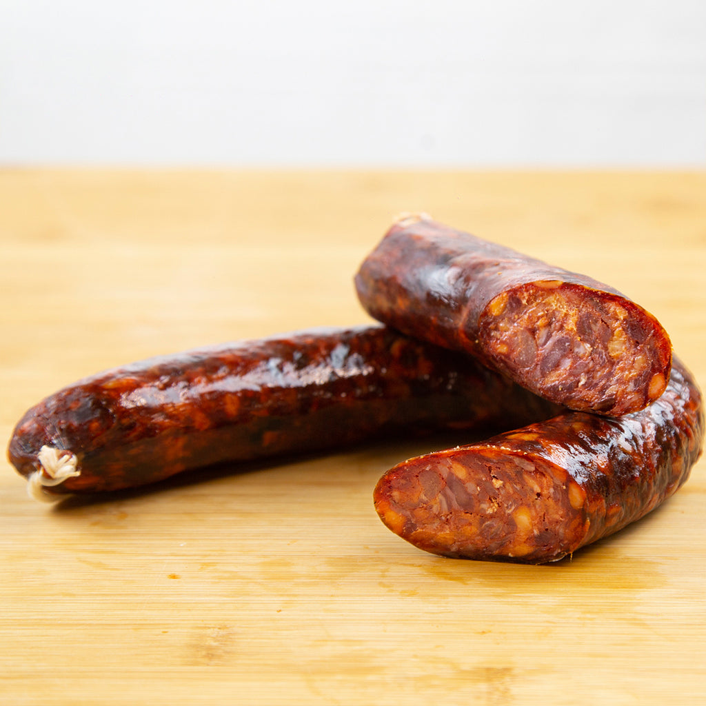 Muestra un chorizo de jabalí en herradura cortado a la mitad, donde se puede apreciar el corte del chorizo. Está sobre una tabla de madera de color marrón claro.