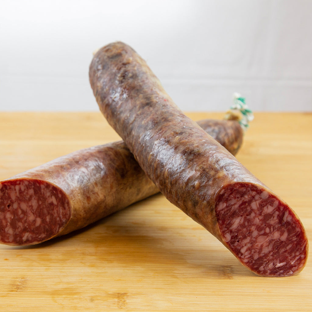 Salchichón ibérico cortado a la mitad, y una mitad puesta sobre la otra de forma que se aprecia la carne y la grasa. El producto está sobre una tabla de madera de color claro. 