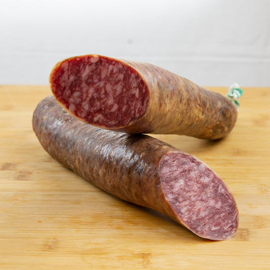 Salchichón ibérico cortado a la mitad, y una mitad puesta sobre la otra de forma que se aprecia la carne y la grasa. El producto está sobre una tabla de madera de color claro.