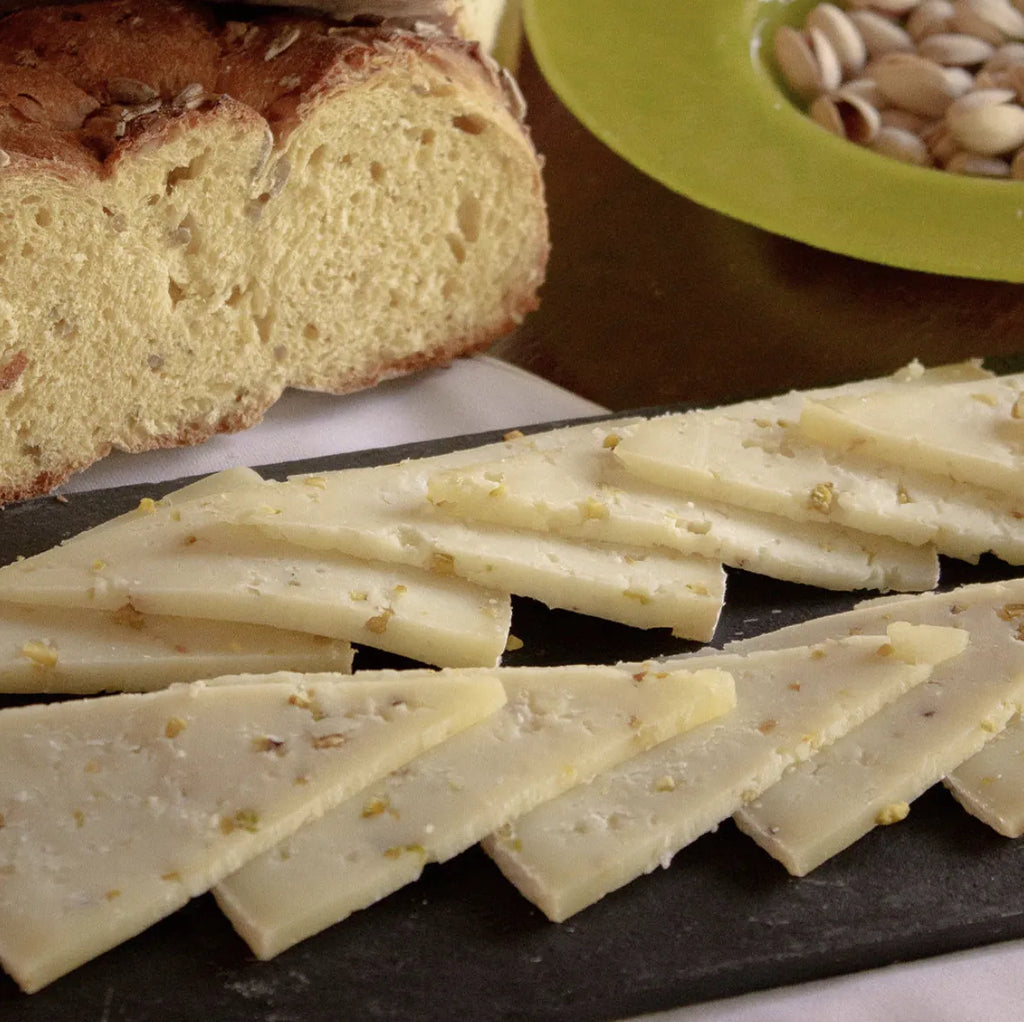 Trozos de pistacho cortado sobre una tabla negra. Al fondo aparece pan y unos pistachos en un plato.