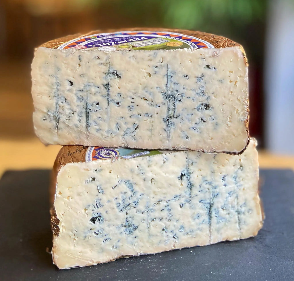 Se muestra un queso azul cortado a la mitad y puesto una parte sobre la otra. El queso aún tiene el papel que lo recubre y el fondo está ligeramente difuminado.
