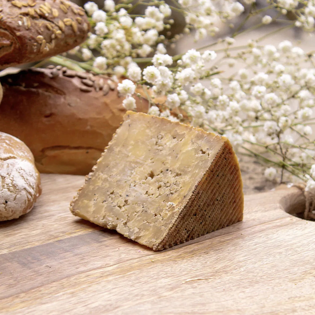 Cuña de queso artesanal de ajo negro sobre mesa de madera. En el fondo aparecen flores blancas y pan.