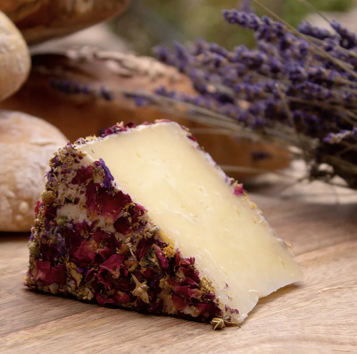 Cuña de queso de oveja con flores en su corteza en primer plano. En segundo plano flores lilas y pan.