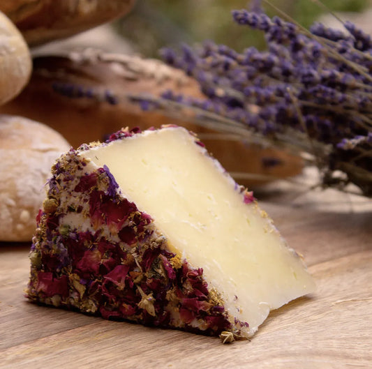 Cuña de queso de oveja con flores en su corteza en primer plano. En segundo plano flores lilas y pan.