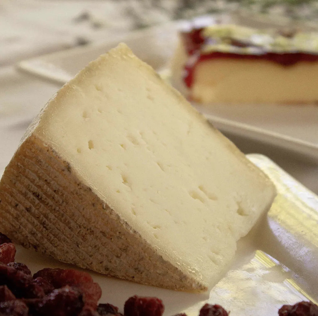 Cuña de queso ligeramente amarillenta. En los márgenes de las fotos se muestran uvas pasas y una tarta de queso al fondo.