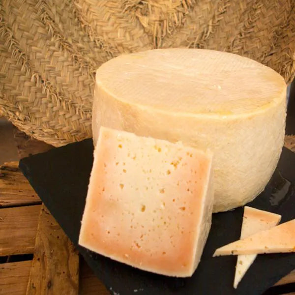 Se muestra un queso entero y una cuña de queso sobre una tabla de color negro.