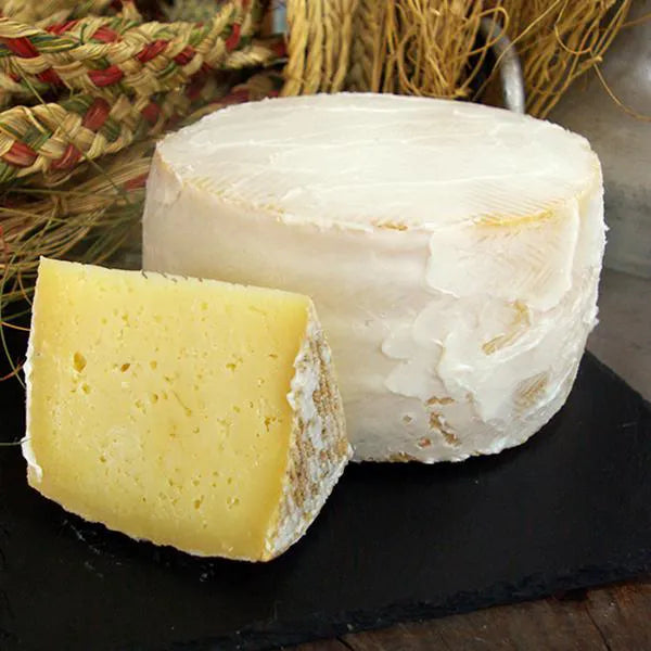 Se muestra un queso entero recubierto de manteca blanca. Además, justo delante se muestra una cuña del queso. Todo sobre una tabla negra.