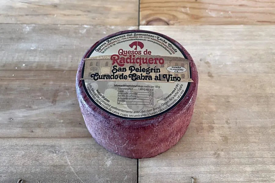 Se muestra un queso entero de corteza rojiza y con una etiqueta en su base superior. El queso está sobre una superficie de madera.
