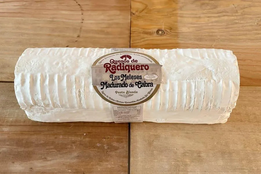 Se muestra un queso alargado de color blanco con una pegatina del productor en el centro. El queso está sobre una superficie de madera.
