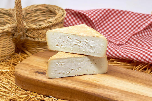 Se muestran 2 cuñas de queso sobre una tabla de madera y al fono dos cestos de paja y un paño de cuadros blanco y rojo.
